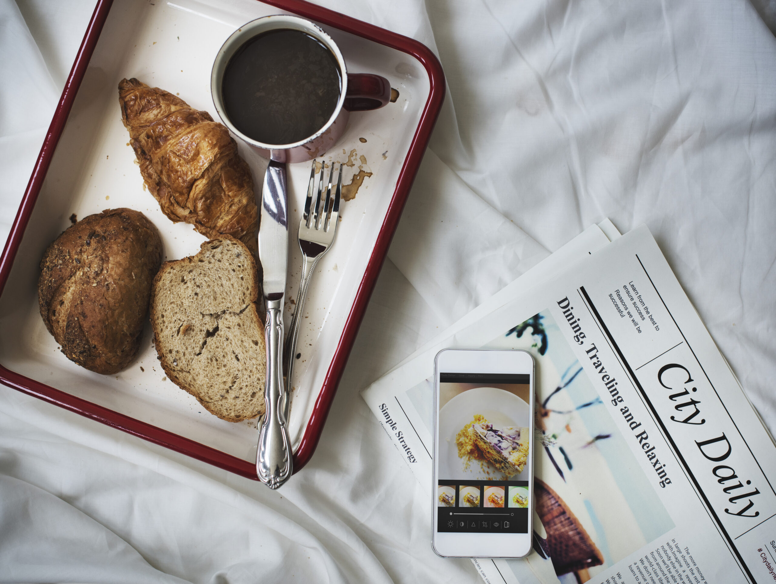 Room service breakfast tray with coffee, croissant, and bread on a bed next to a smartphone and a newspaper titled "City Daily."